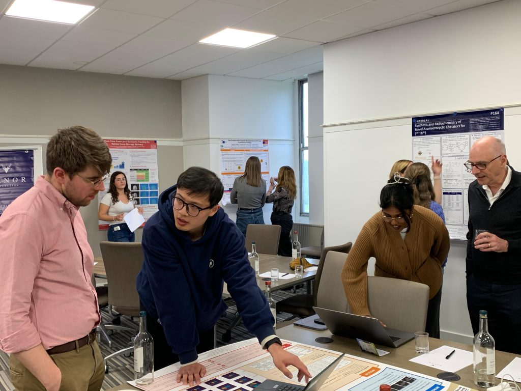 Attendees of the Medical Imaging CDT event interacting during a poster session in a conference room, with laptops, papers, and scientific posters displayed on the walls